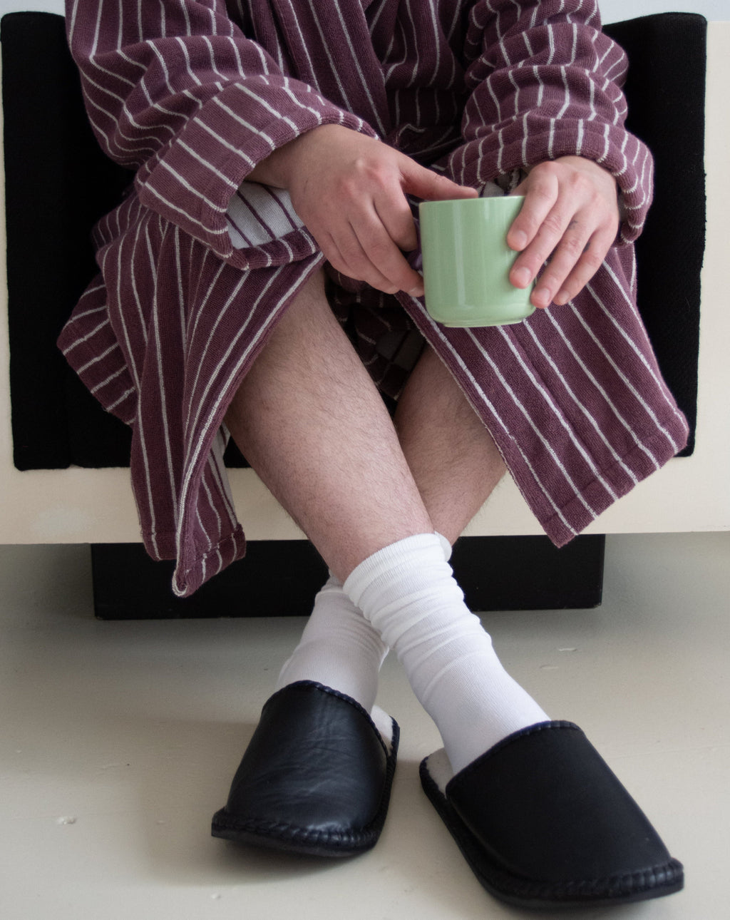 Person wearing a striped robe, white socks, and black slippers holding a green mug.