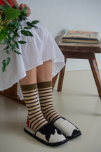 Person wearing striped socks and black and white slippers, sitting on a stool with a plant and books in the background.