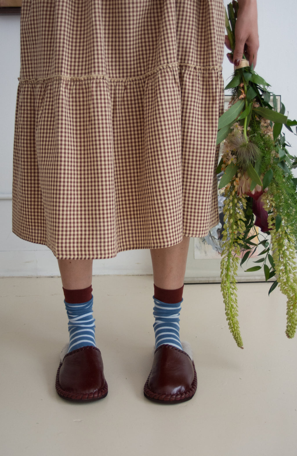 Person wearing a checkered dress, striped socks, and brown shoes holding greenery indoors.