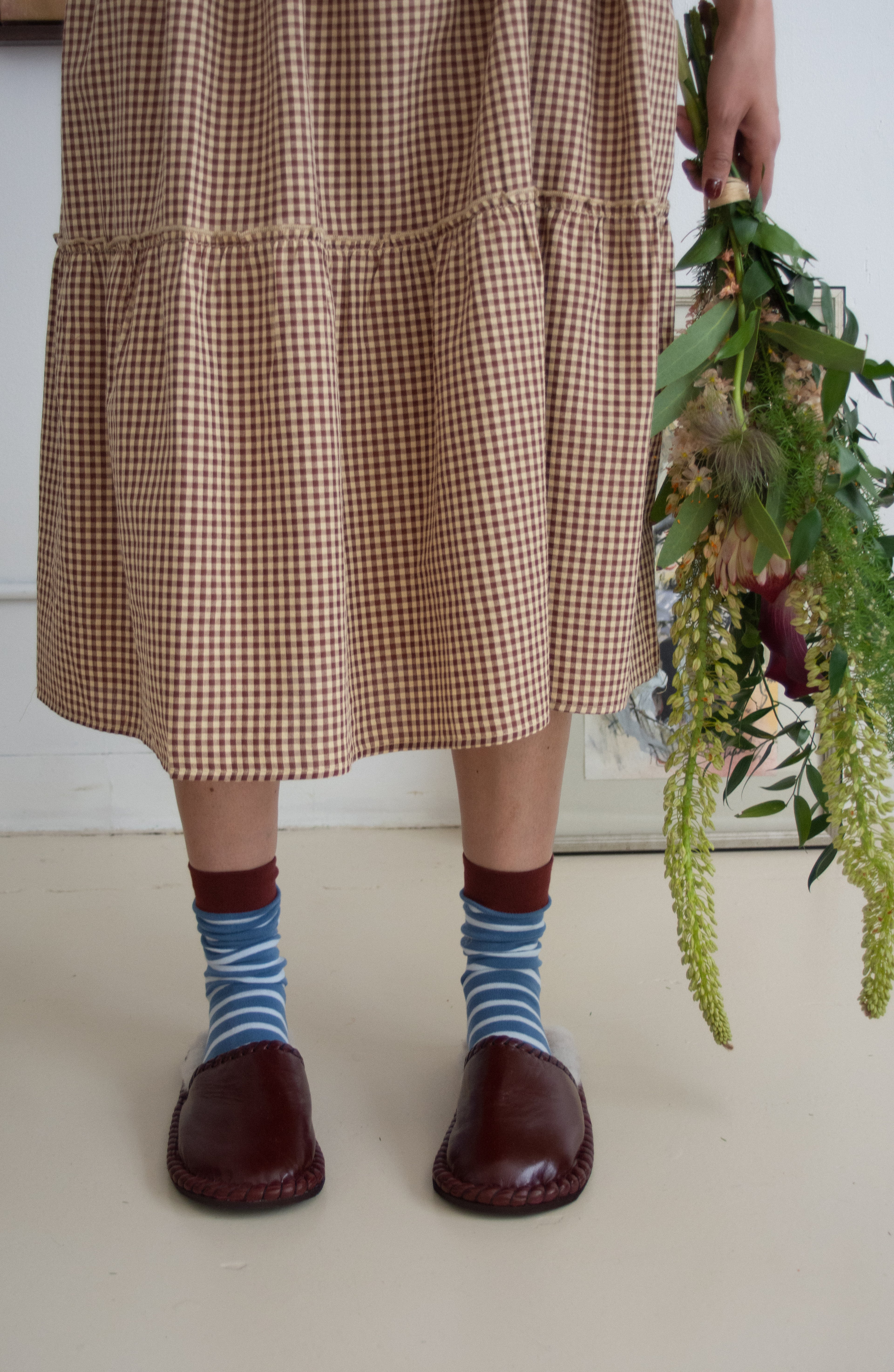 Person wearing a checkered dress, striped socks, and brown shoes holding greenery indoors.
